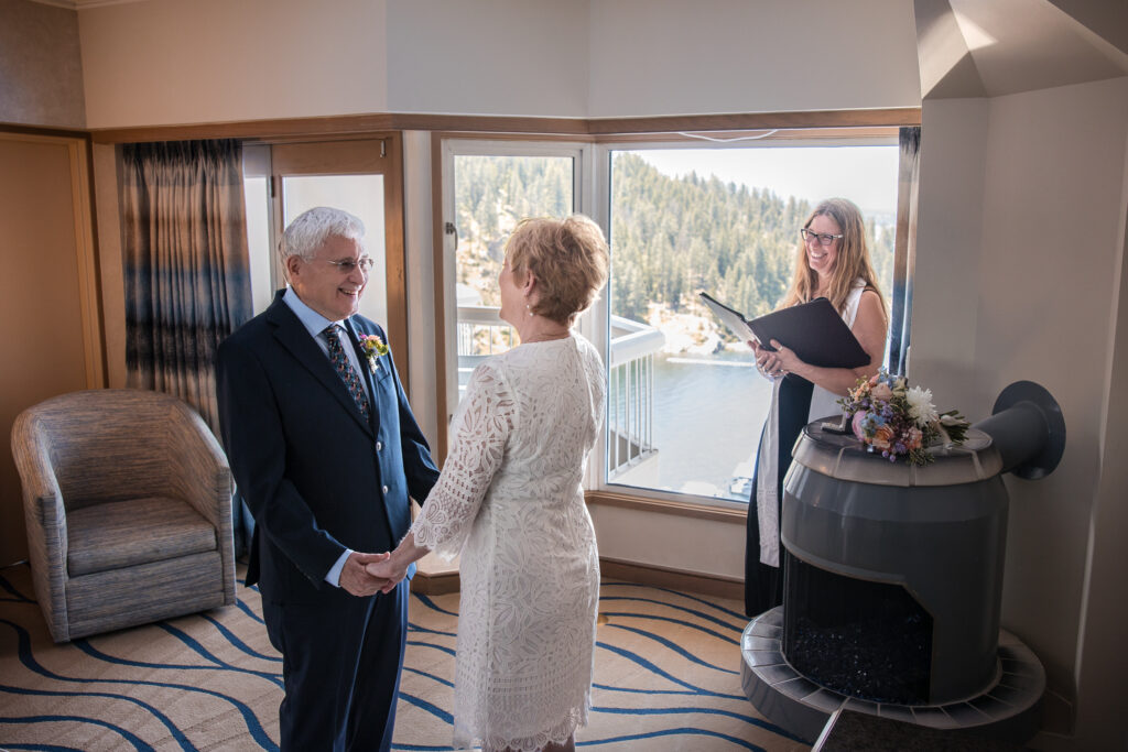 Groom smiling at bride while officiant leads vows in suite with lake view