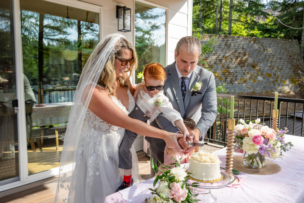 Couple cutting cake outdoors on deck with Coeur d’Alene Lake behind them.holding son
