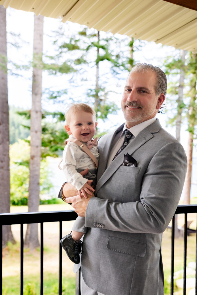 Groom in a grey suit holding a baby dressed in pink suspenders and bow ties on the deck.