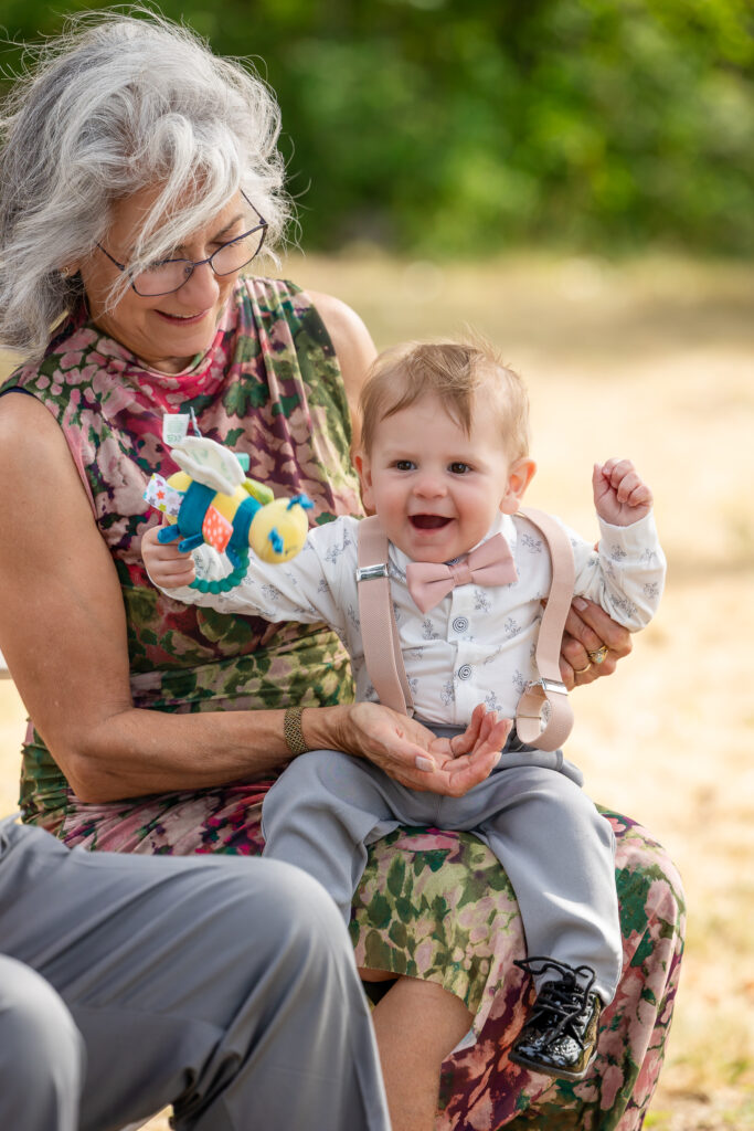 Baby sitting on grandma's lap, smiling, wearing a pink bow tie and suspenders